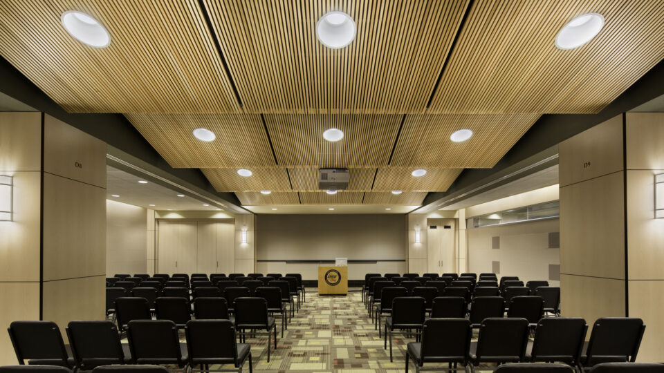 Empty conference room with rows of black chairs, a podium, and a wood slat ceiling with recessed lights.