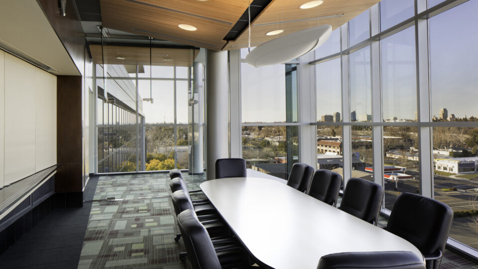 Modern conference room with a long white table, black chairs, floor-to-ceiling windows, and city views.