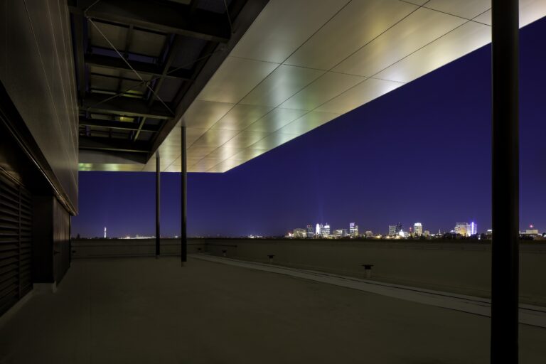 Modern rooftop terrace at dusk with a glowing metal canopy and a distant city skyline under a deep blue sky