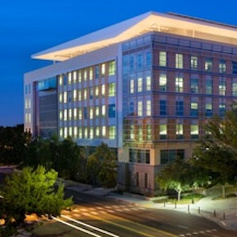 Modern multi-story office building illuminated at dusk, with glass and brick exterior above a tree-lined street