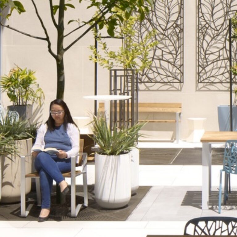 Woman sitting in a chair and looking at her phone in a bright indoor courtyard with potted plants and tree decor