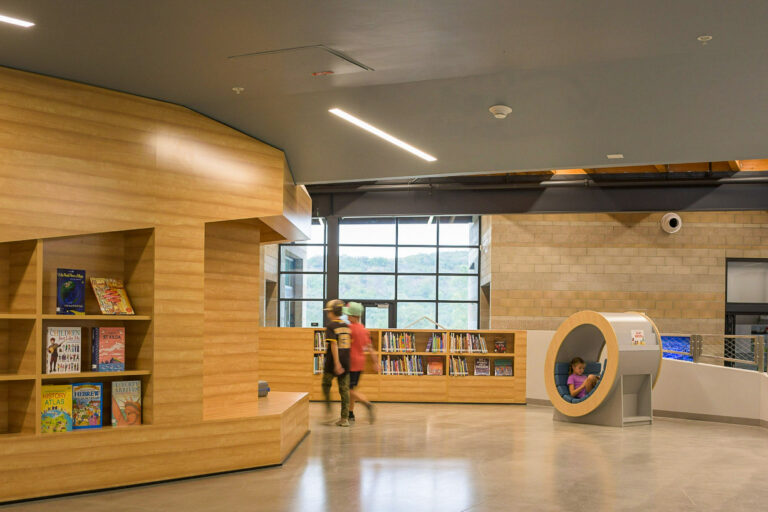 Children’s library interior with wooden shelves, two kids walking, and a girl reading in a round nook seat.