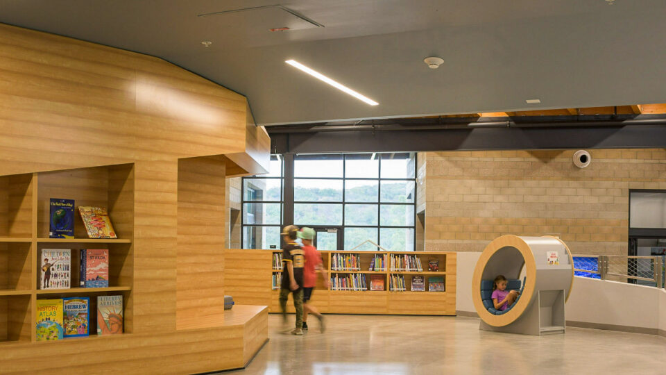 Children’s library interior with wooden shelves, two kids walking, and a girl reading in a round nook seat.