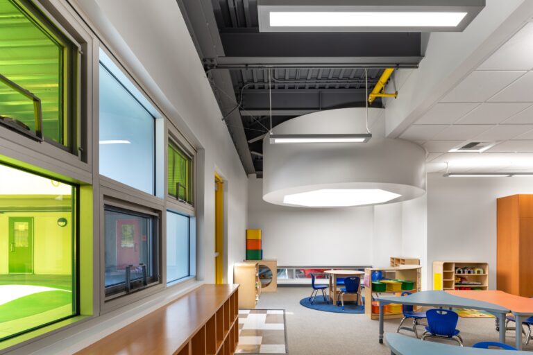 Bright modern classroom with blue child-sized chairs, curved tables, wood shelving, and large green-tinted windows.