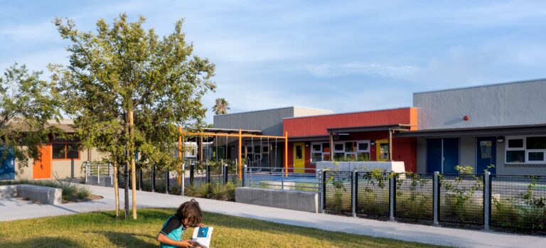 Child reading on a grassy lawn in front of a colorful modern school building with trees and a fenced walkway