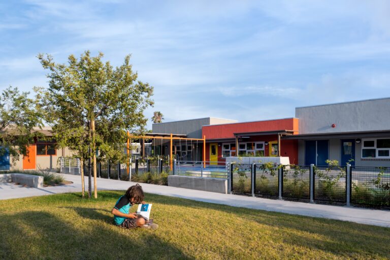 Child reading on a grassy lawn in front of a colorful modern school building with trees and a fenced walkway