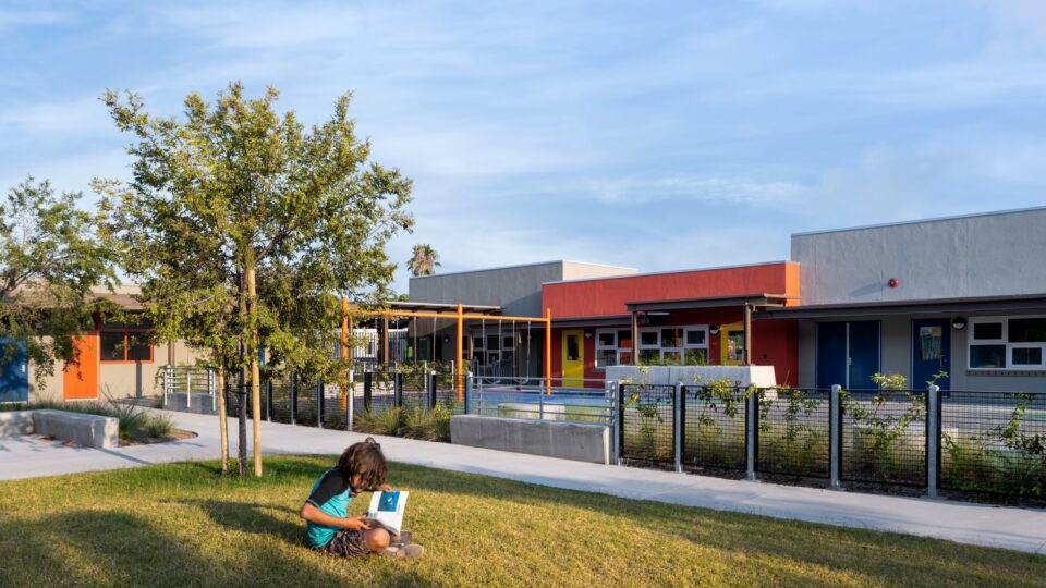Child reading on a grassy lawn in front of a colorful modern school building with trees and a fenced walkway