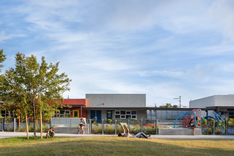 Children reading and playing on a grassy lawn outside a modern school with a colorful playground under a wide blue sky