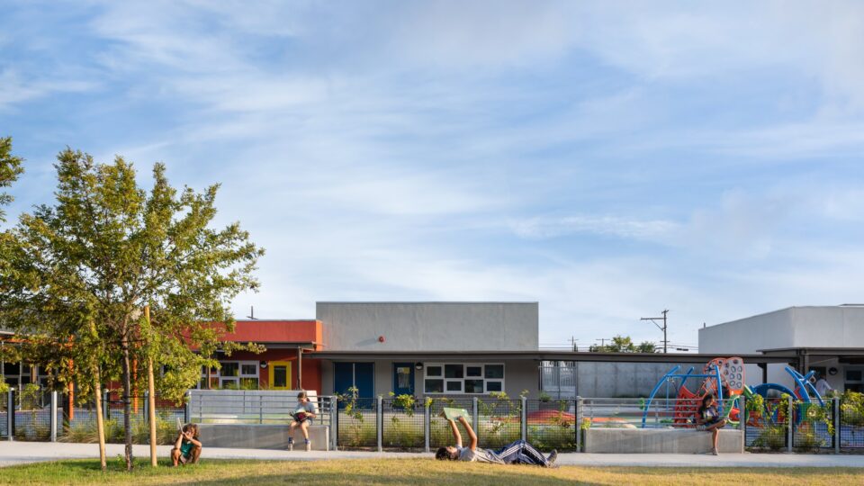Children reading and playing on a grassy lawn outside a modern school with a colorful playground under a wide blue sky