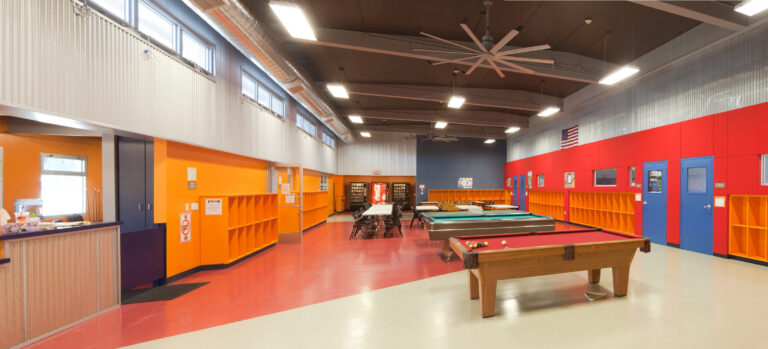 Bright recreation room with pool tables, tables and chairs, orange cubbies, and high ceilings with large fans.