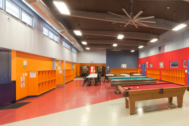 Bright recreation room with pool tables, tables and chairs, orange cubbies, and high ceilings with large fans.