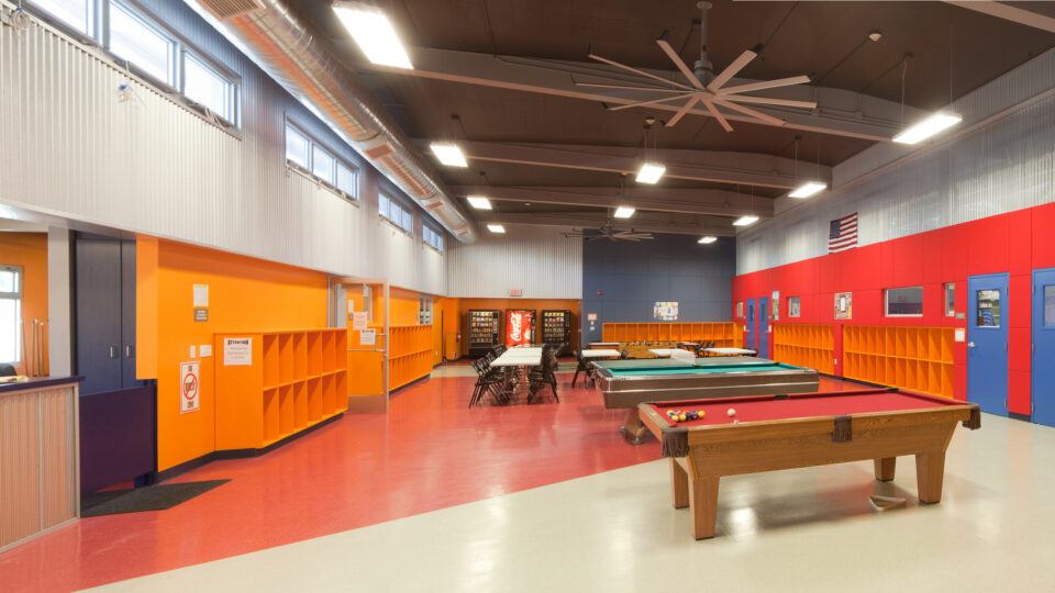 Bright recreation room with pool tables, tables and chairs, orange cubbies, and high ceilings with large fans.