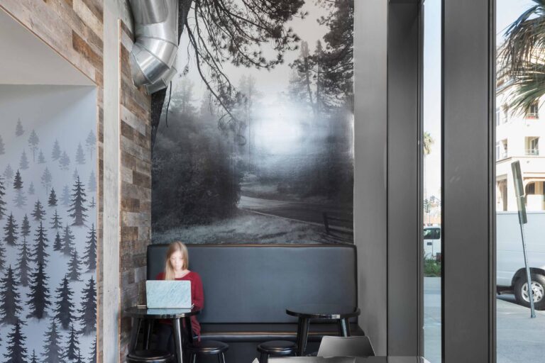 Modern café interior with tall forest mural, exposed ductwork, and a person seated at a booth using a laptop