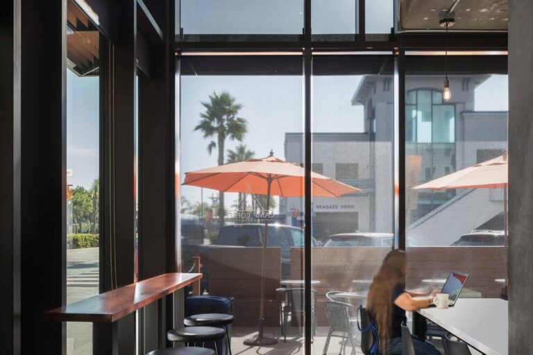 Modern cafe interior with tall windows, bar stools, and a woman working at a table beside a sunny patio.