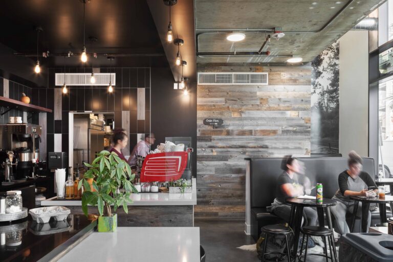 Modern coffee shop interior with pendant lights, espresso bar, and two blurred customers seated by tall windows.