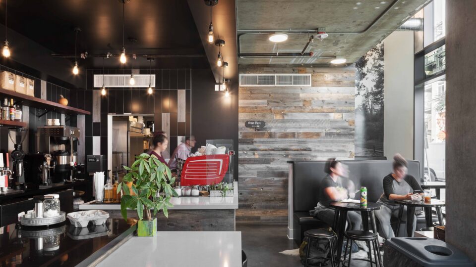 Modern coffee shop interior with pendant lights, espresso bar, and two blurred customers seated by tall windows.