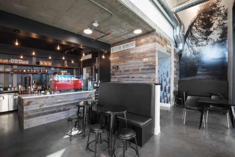 Modern coffee shop interior with a red espresso machine, rustic wood walls, black booth seating, and forest mural.