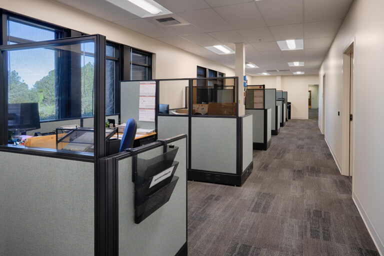 Office hallway lined with gray cubicles and large windows, with desks and chairs inside the workstations
