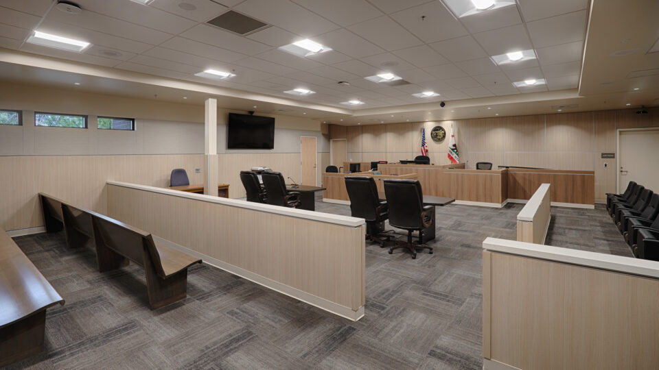 Empty modern courtroom with judge’s bench, witness stand, counsel tables, benches, and U.S. and California flags