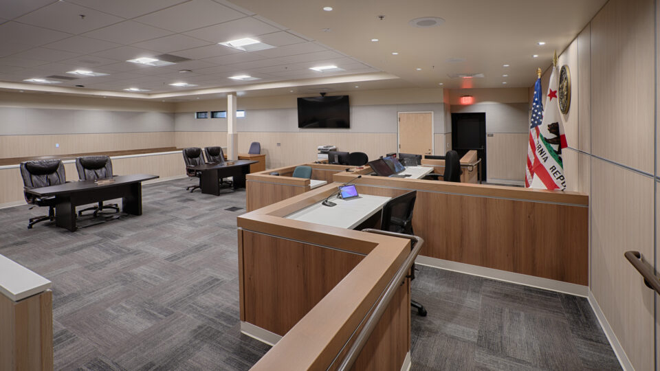 Empty courtroom interior with judge’s bench, counsel tables, witness stand, and U.S. and California flags.