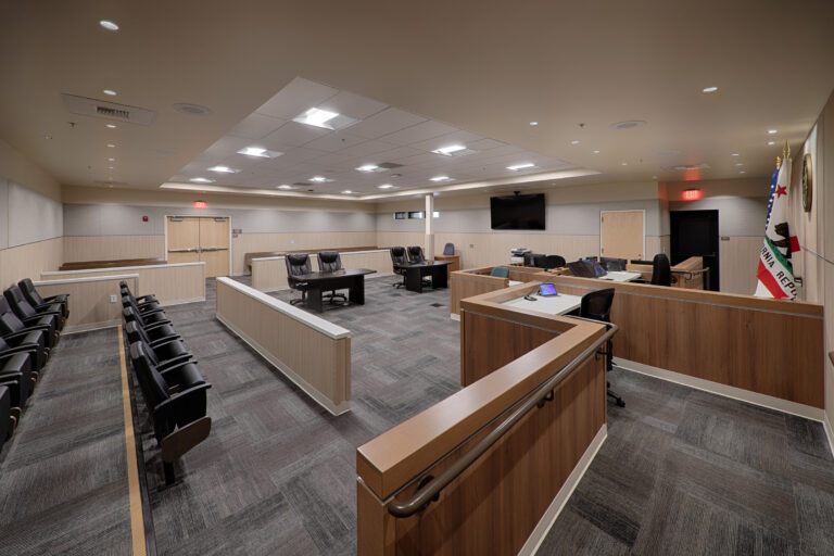 Empty courtroom with wood-paneled benches, judge’s desks, spectator seating, and a California Republic flag.
