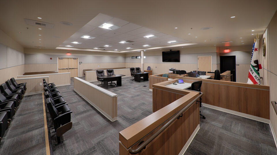 Empty courtroom with wood-paneled benches, judge’s desks, spectator seating, and a California Republic flag.