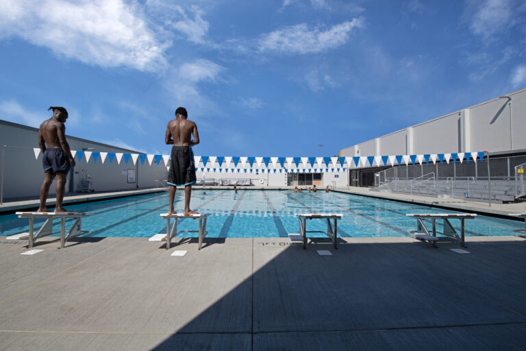Two swimmers stand on starting blocks beside an outdoor lap pool under a bright blue sky.