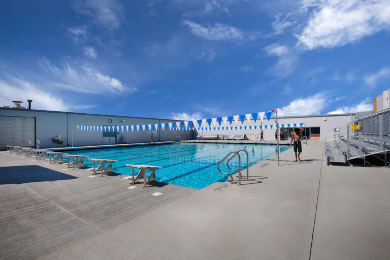 Outdoor lap pool with starting blocks, blue-and-white lane flags, and a few swimmers under a bright blue sky.