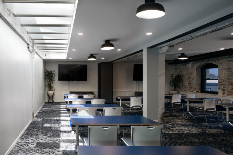 Modern classroom with blue tables, white chairs, exposed brick, skylight, and wall-mounted screens.