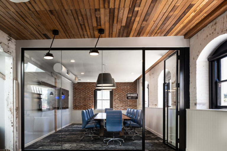 Glass-walled conference room with blue chairs, exposed brick walls, and a wood plank ceiling in a modern office.