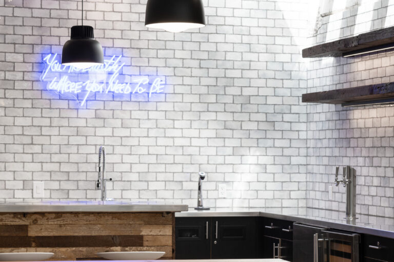 Modern kitchen with white brick walls, black pendant lights, wooden table and stools, and a blue neon sign.