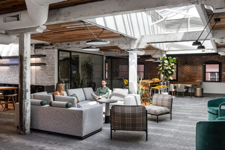 Modern industrial office lounge with skylights, exposed beams, and two people talking on gray sofas.