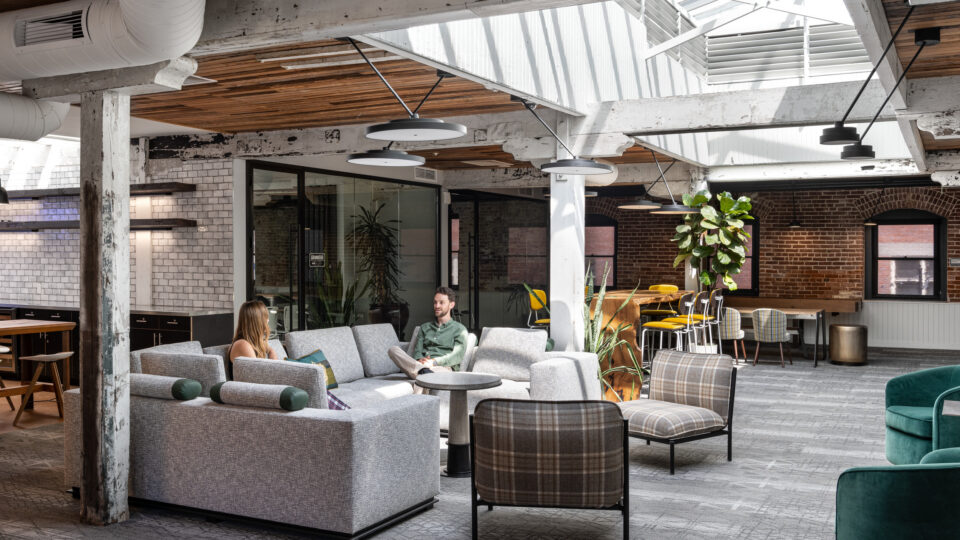 Modern industrial office lounge with skylights, exposed beams, and two people talking on gray sofas.