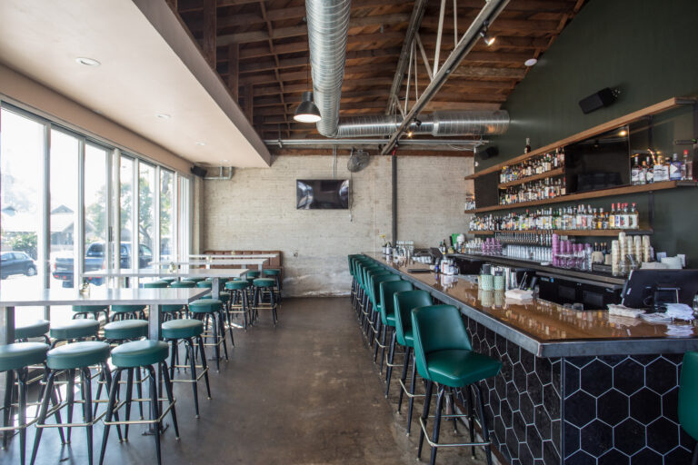 Modern bar interior with green stools, high-top tables, exposed ductwork, and shelves of liquor bottles.