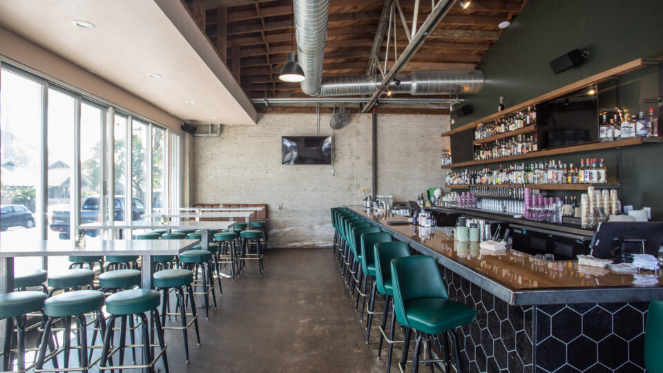 Modern bar interior with green stools, high-top tables, exposed ductwork, and shelves of liquor bottles.