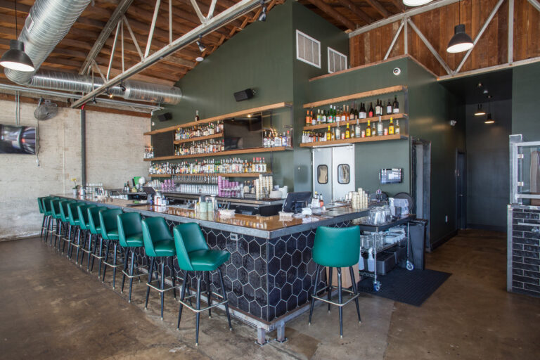 Industrial-style bar interior with green stools, a long tiled counter, and shelves of liquor beneath exposed ductwork.