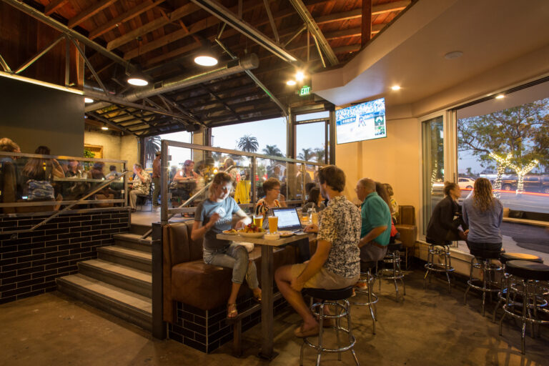 Busy modern bar and restaurant interior with groups dining, large open windows, and a TV showing a baseball game.