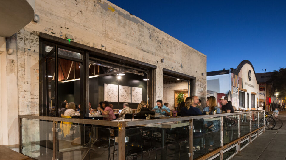People dining on a restaurant patio outside a weathered building at dusk on a city street