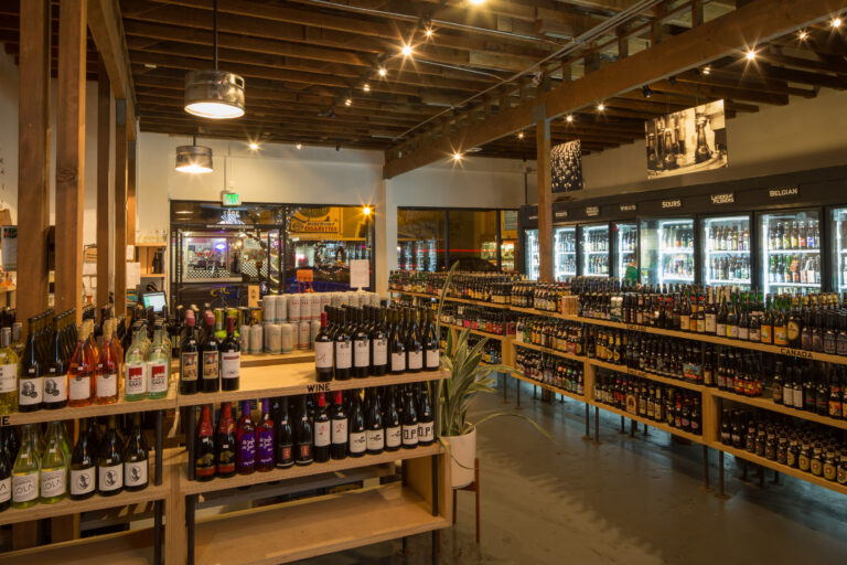 Interior of a bottle shop with wine displays, refrigerated craft beers, and wooden beams under warm lighting