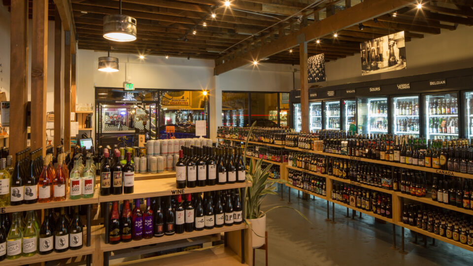 Interior of a bottle shop with wine displays, refrigerated craft beers, and wooden beams under warm lighting