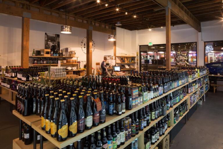 Interior of a craft beer shop with long shelves packed with bottled beers and a clerk at the counter.