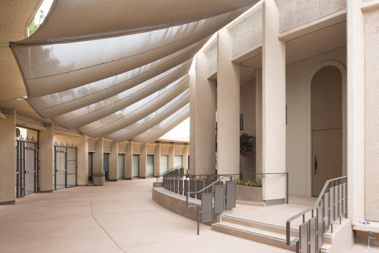 Covered outdoor corridor with large fabric shade sails, stucco columns, and steps with metal railings to an entrance
