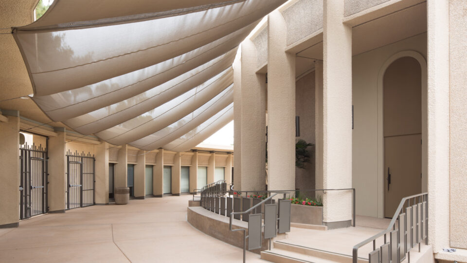 Covered outdoor corridor with large fabric shade sails, stucco columns, and steps with metal railings to an entrance
