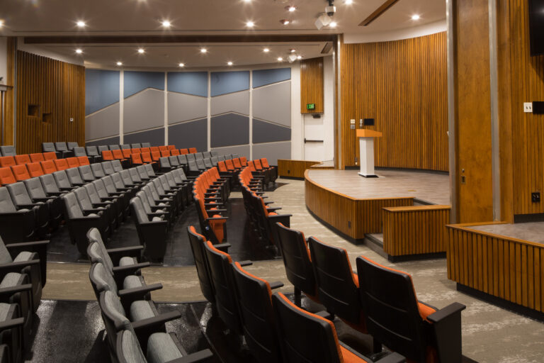 Empty auditorium with curved rows of gray and orange seats facing a small stage and podium.