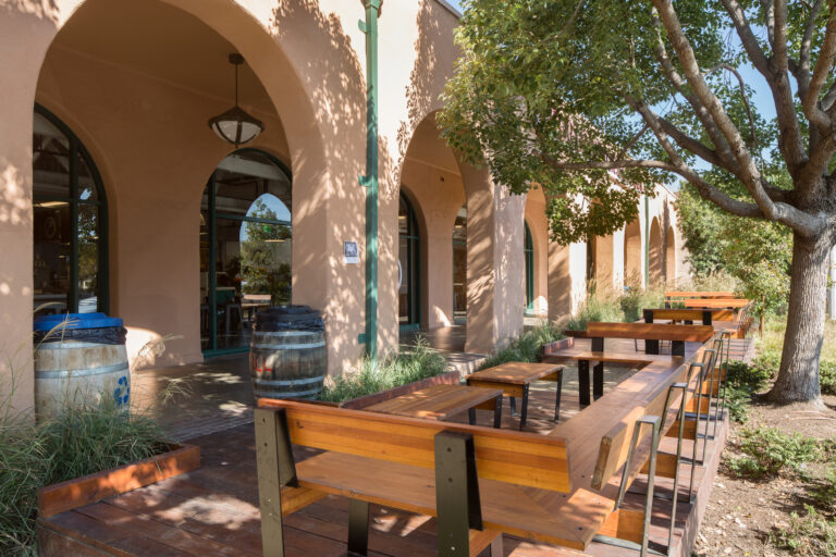 Outdoor wooden bench seating beside an arched stucco building, shaded by trees and surrounded by landscaping.