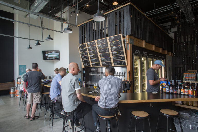 People sitting and standing at a modern brewery taproom bar beneath large beer menu boards and hanging lights