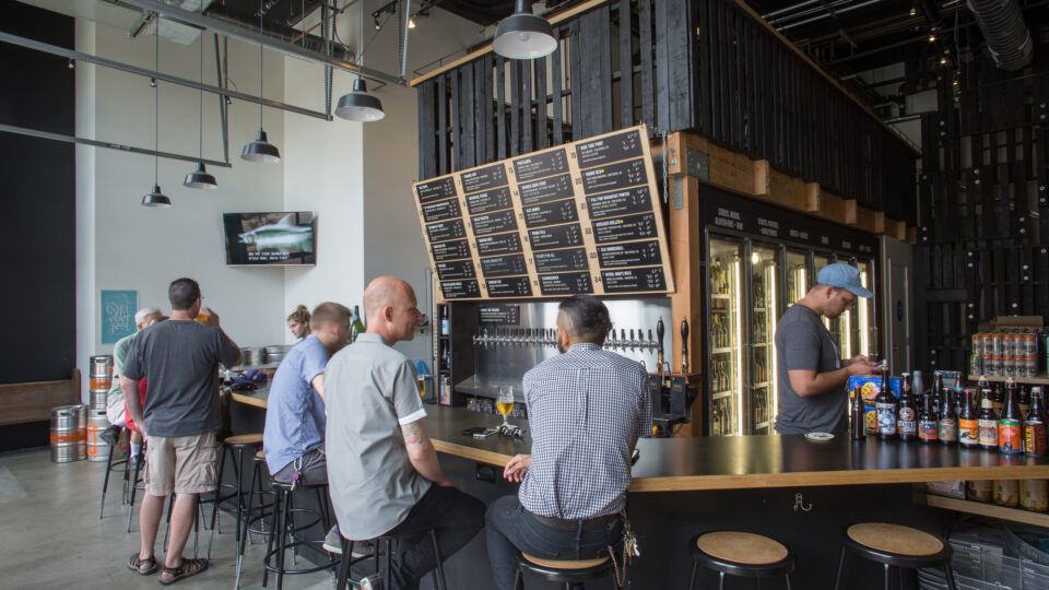 People sitting and standing at a modern brewery taproom bar beneath large beer menu boards and hanging lights