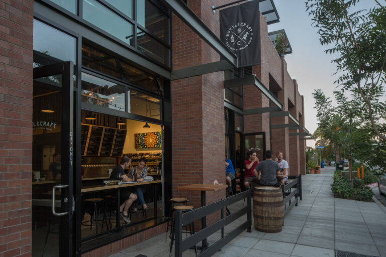 Brick storefront of Bottlecraft beer shop and tasting room with open windows and people seated inside and outside.