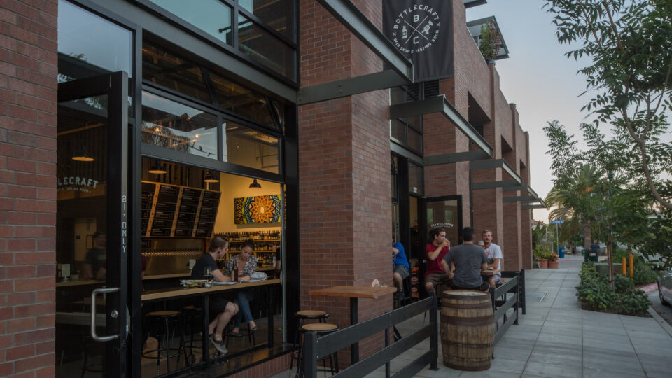 Brick storefront of Bottlecraft beer shop and tasting room with open windows and people seated inside and outside.