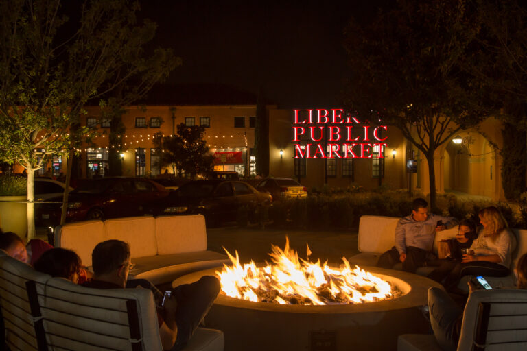 People relax around an outdoor fire pit at night in front of the glowing Liberty Public Market sign.